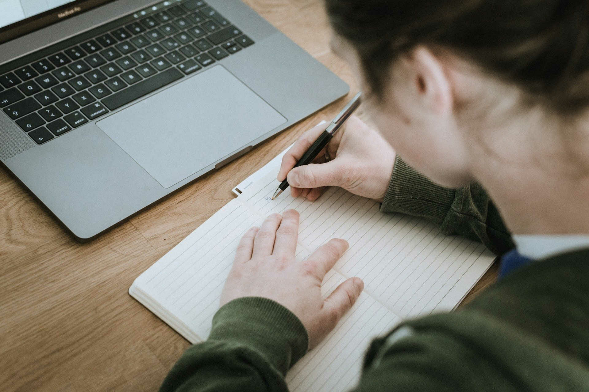 Student writing in notepad with laptop in front of her